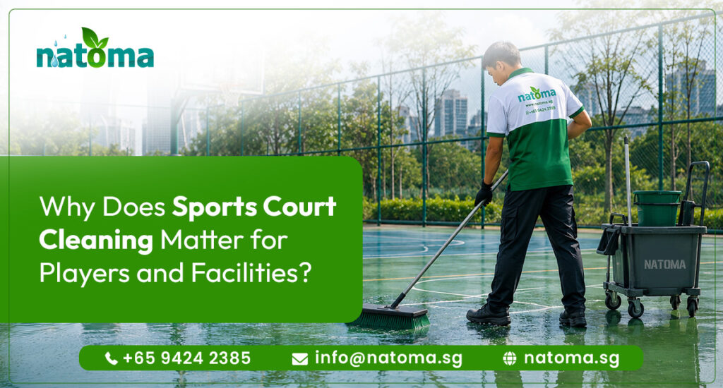 A professional cleaner wearing a white and green uniform shirt, dark pants, and gloves uses a long-handled squeegee to clear water or clean the surface of an outdoor sports court.